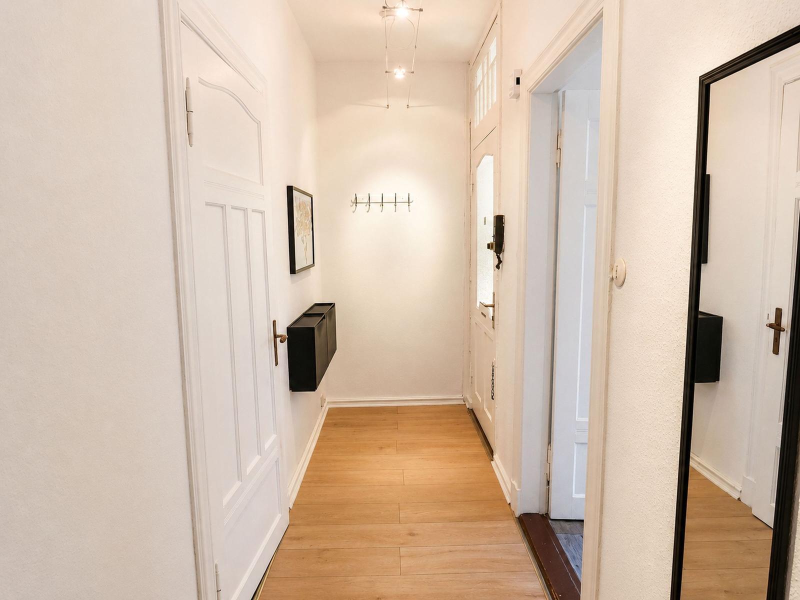 Flur mit Holzboden, weißen Türen und Wandgarderobe. Spiegel reflektiert den Raum.