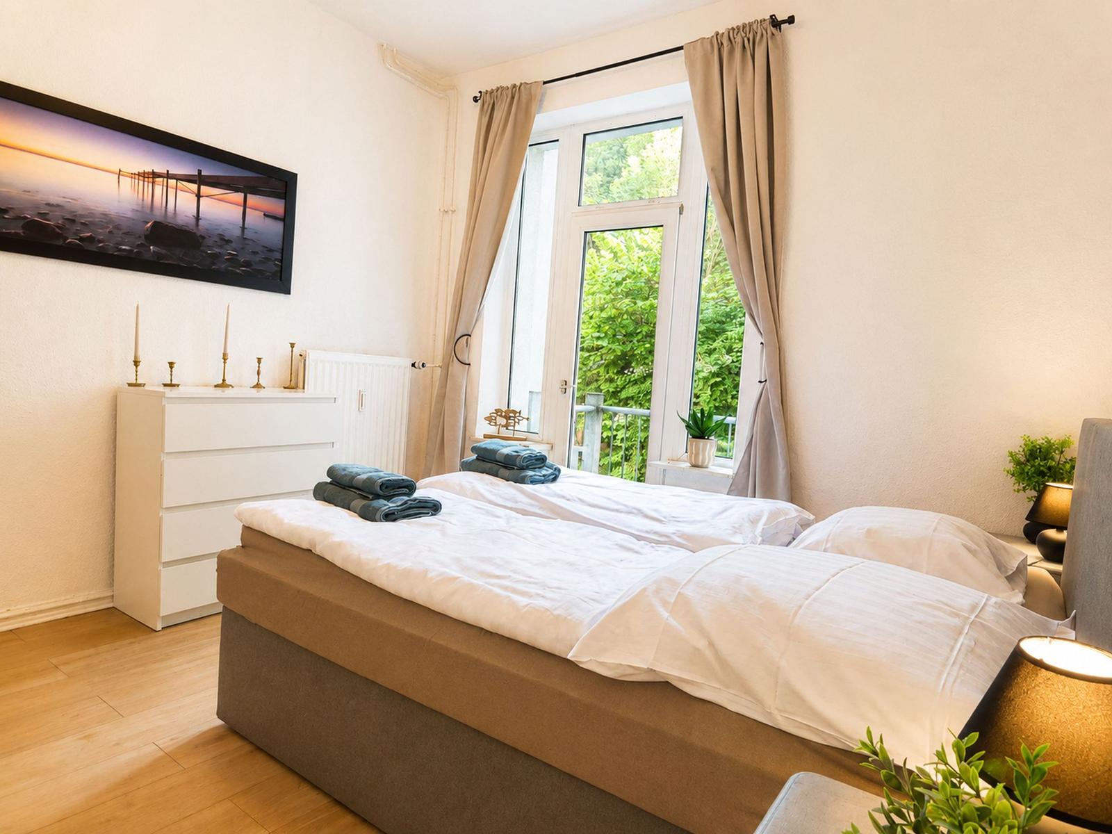 Schlafzimmer mit Doppelbett, Fenster mit Blick auf Garten und weiße Kommode mit Kerzen.