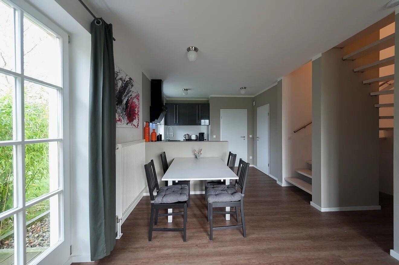 Essbereich mit Tisch und Stühlen, Küche im Hintergrund, große Fenster und Treppe zur oberen Etage.