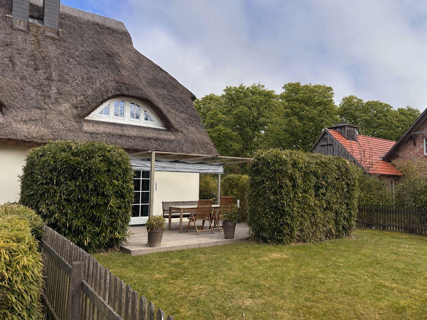 Haus mit Strohdach, Terrasse mit Tisch und Stühlen, umgeben von grünen Hecken.