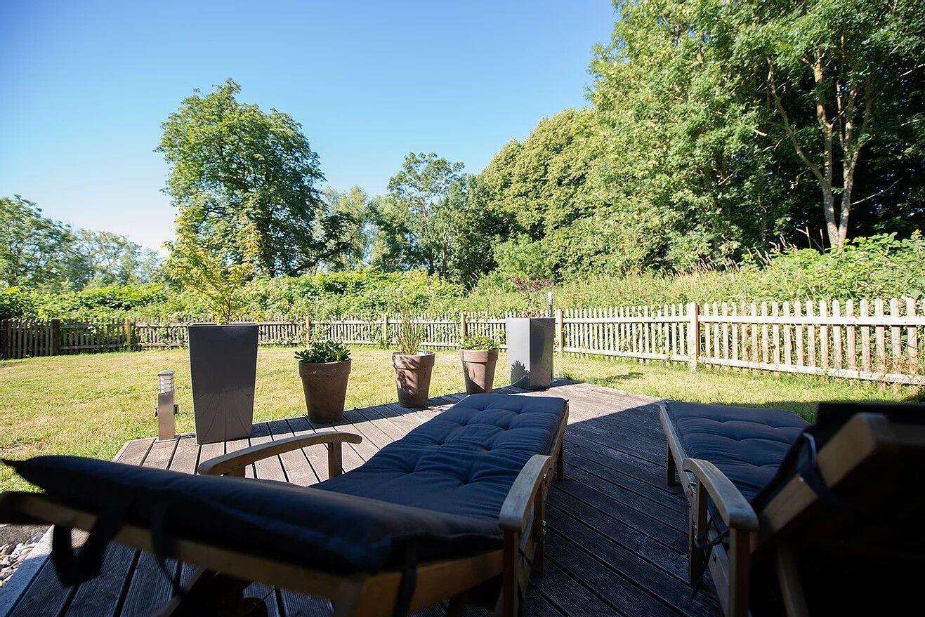 Terrasse mit Liegestühlen, Pflanzen und Holzgeländer. Grünflächen und Bäume im Hintergrund.