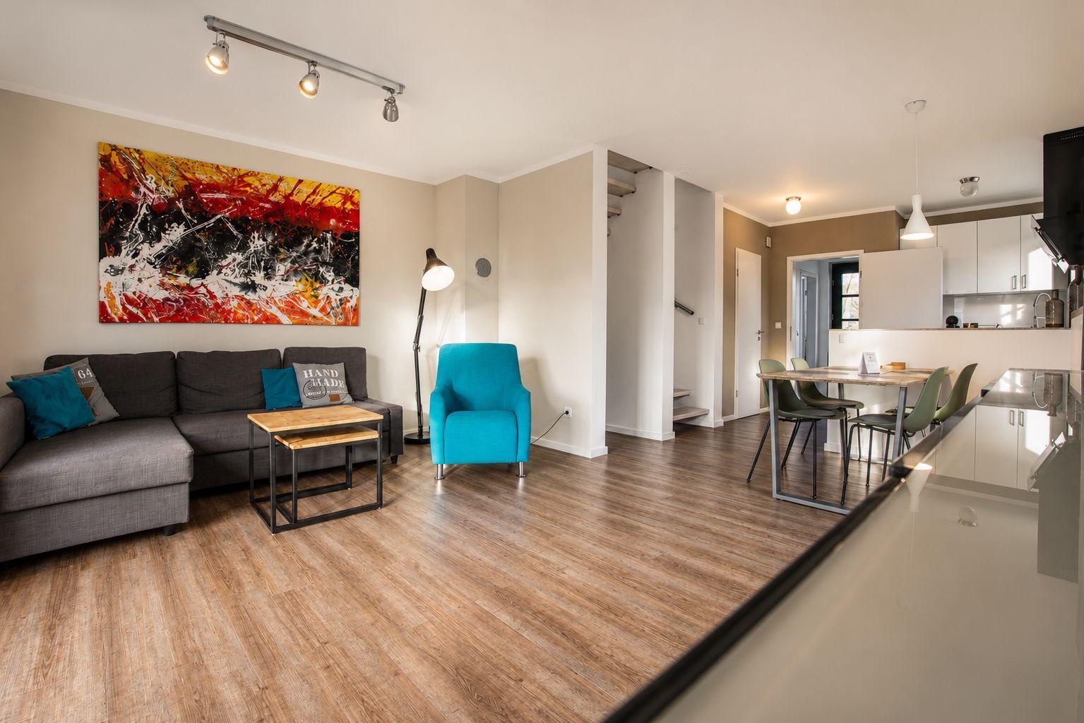 Wohnzimmer mit Sofa, Sessel und Küchenbereich. Holzboden und Wandbild sichtbar.