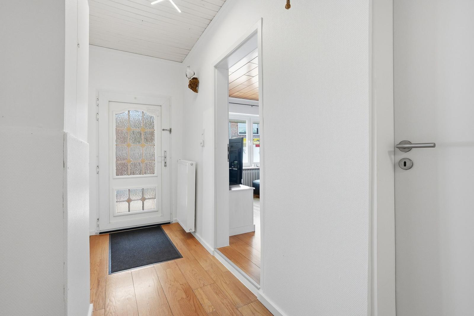 Hausflur mit Holzboden, weißen Wänden und Tür zum Wohnzimmer.