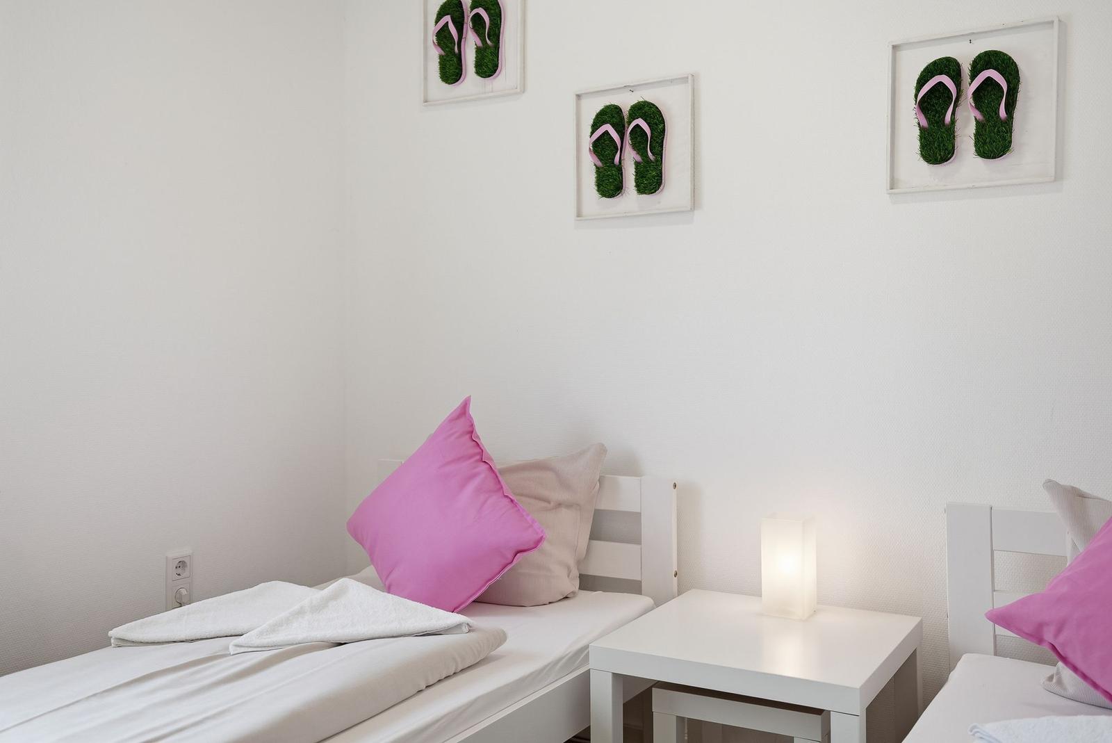 Zwei Betten mit Kissen und einer Nachttischlampe. An der Wand hängen drei Bilder mit Flip-Flops.
