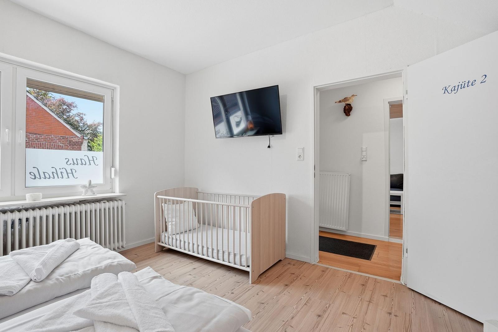 Zimmer mit Babybett, TV und Fenster. Tür führt in Wohnbereich.