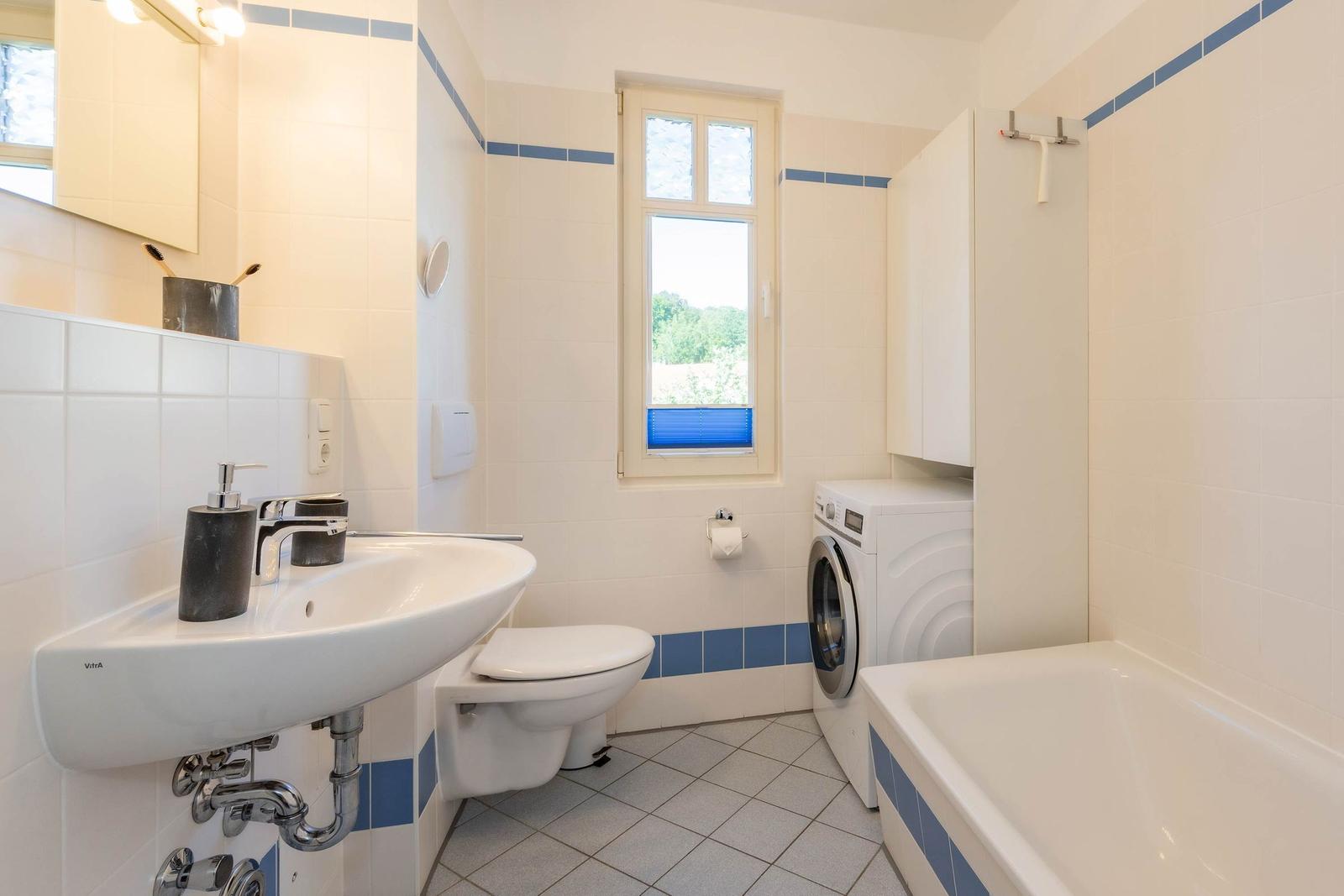 Badezimmer mit Waschmaschine, Badewanne und Toilette. Fenster mit blauen Jalousien.