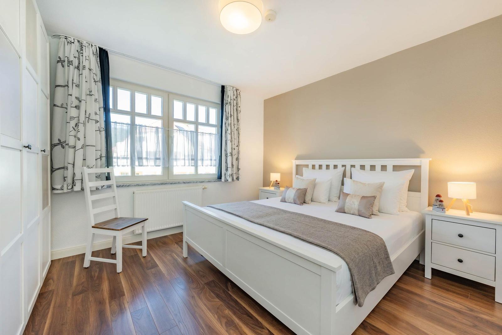 Schlafzimmer mit weißem Bett, Nachttischen und Holzboden. Fenster mit Vorhängen.