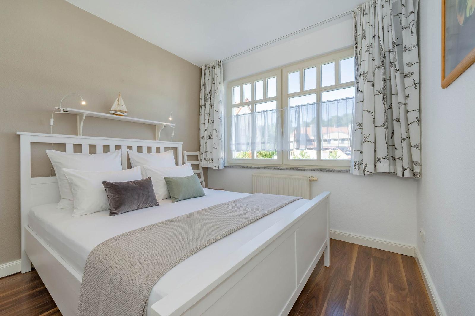 Schlafzimmer mit weißem Bett, Fenster mit Vorhängen und Holzboden.