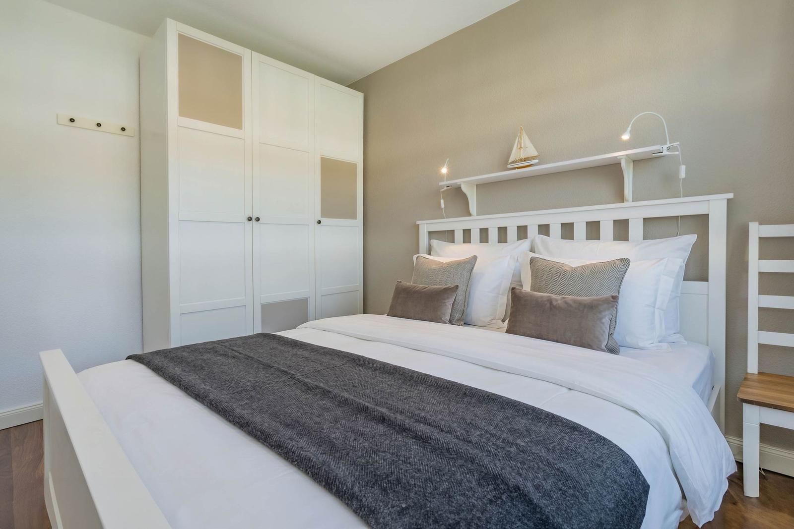 Schlafzimmer mit weißem Bett, Kleiderschrank und Nachttisch. Auf dem Kopfteil steht ein Segelboot.