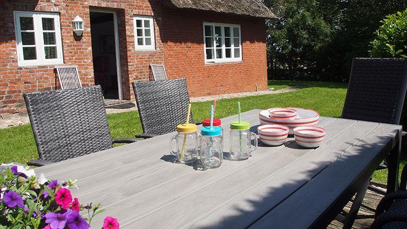 Holztisch mit Stühlen, Tellern und Getränkegläsern im Garten vor einem Backsteinhaus.
