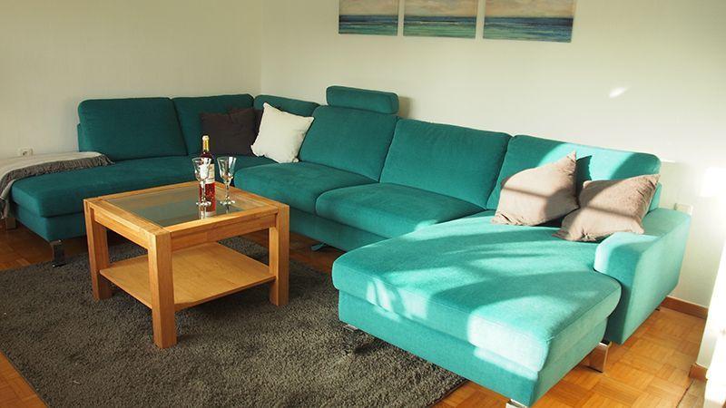 Ein Wohnzimmer mit einem türkisfarbenen Ecksofa, einem Holztisch und Wandbildern.