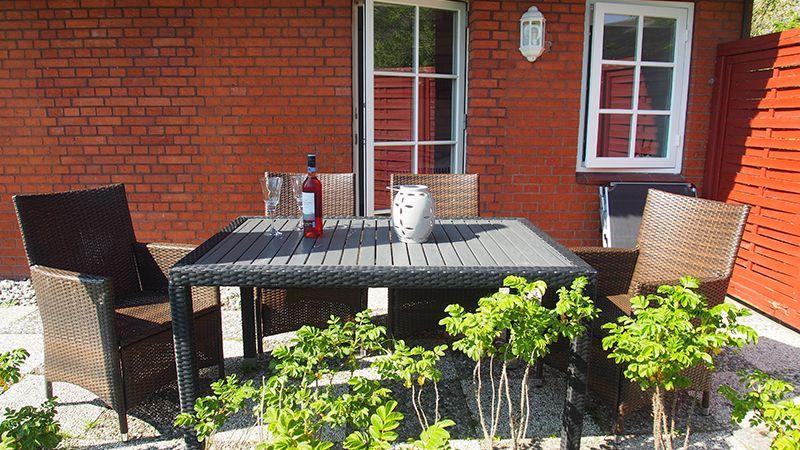 Terrasse mit Tisch, Stühlen und einer Flasche. Hintergrund: rote Backsteinwand und Fenster.