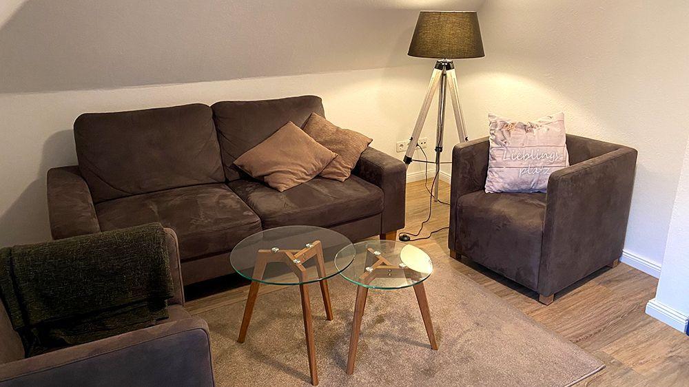 Wohnzimmer mit braunem Sofa, Sessel, Glas-Tisch und Stehlampe.