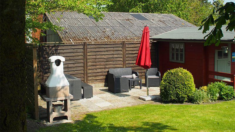 Roter Gartenhäuschen mit Terrasse, Grill und Sitzmöbel unter rotem Sonnenschirm.