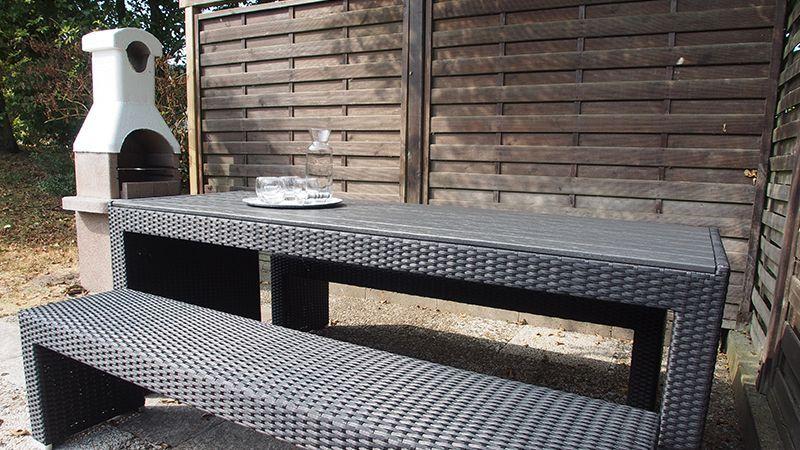 Außenbereich mit Tisch, Bank und Grill. Holzwand im Hintergrund.