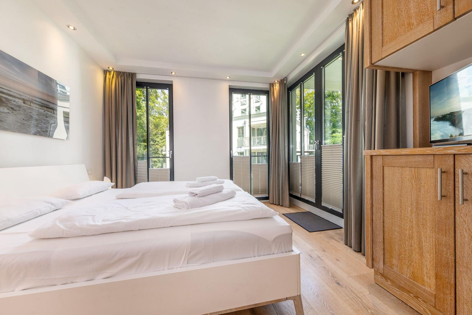 Schlafzimmer mit Doppelbett, Holzboden und großen Fenstern mit Blick auf Balkon.