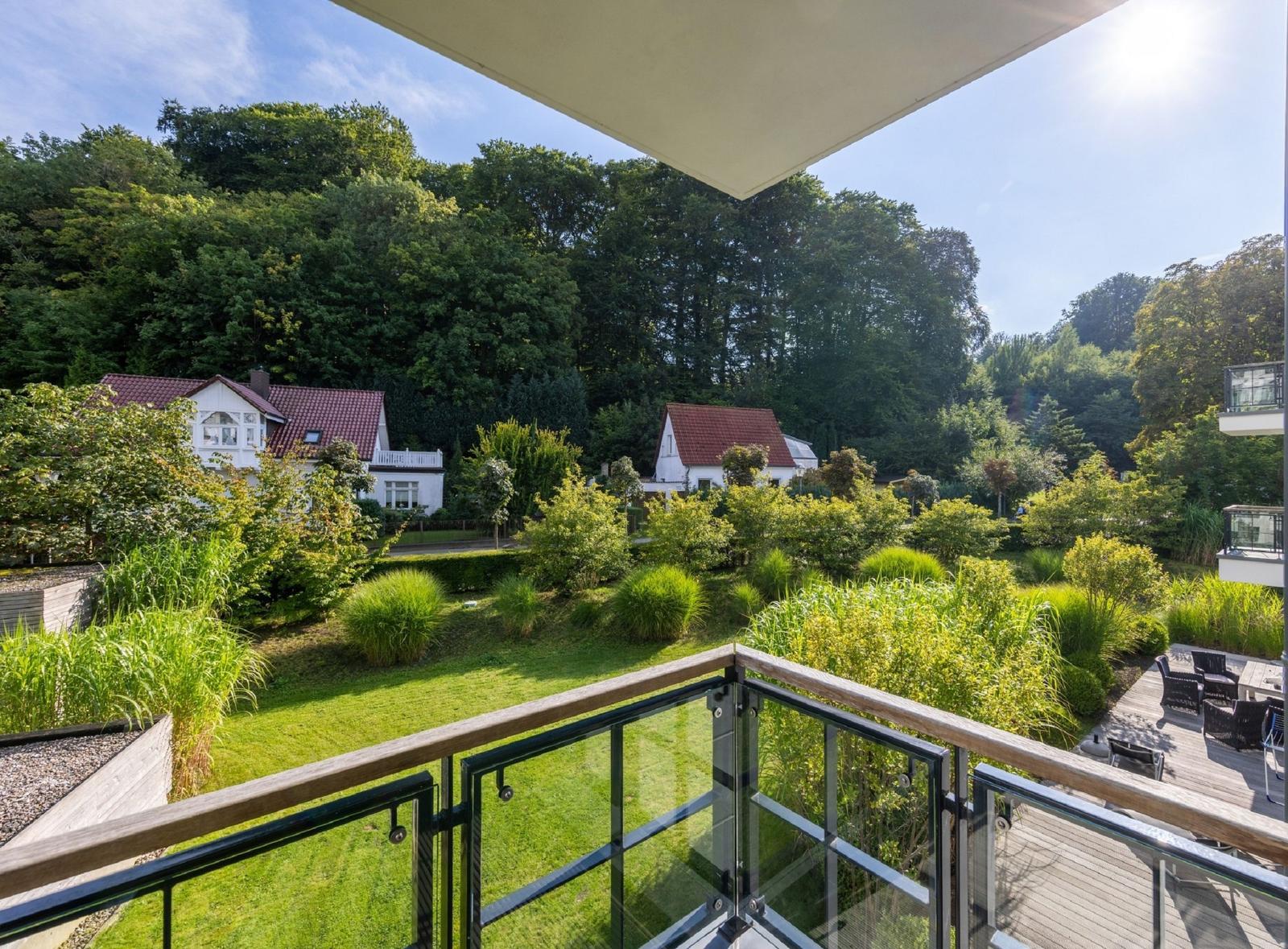 Balkon mit Glasgeländer bietet Blick auf grünen Garten und Häuser im Hintergrund.
