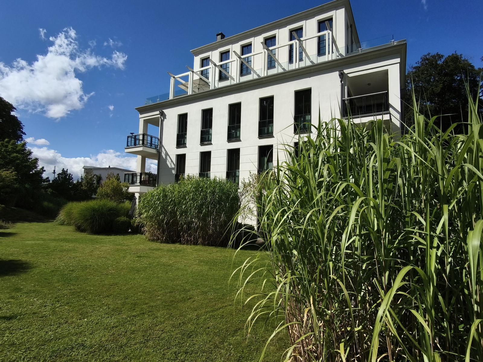 Modernes weißes Gebäude mit Balkonen und grünem Garten unter blauem Himmel.