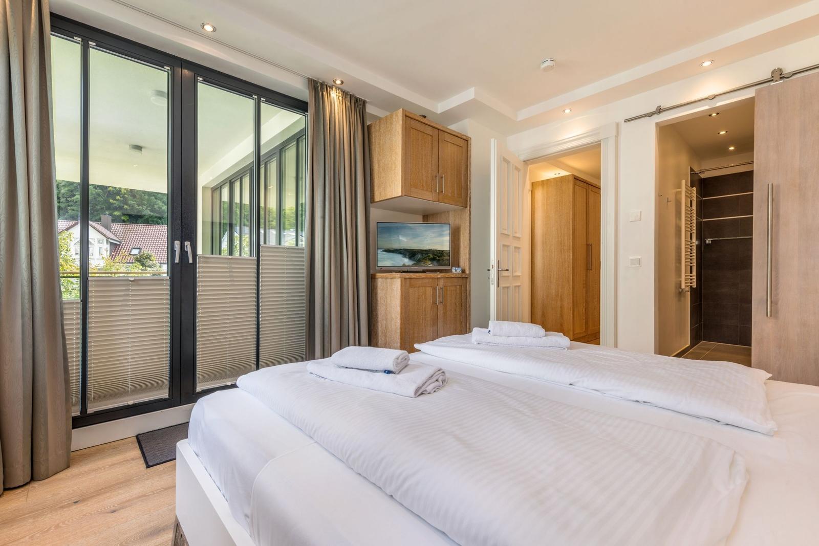 Schlafzimmer mit Doppelbett, TV und Balkonansicht. Dusche und Schrank im Anschluss.