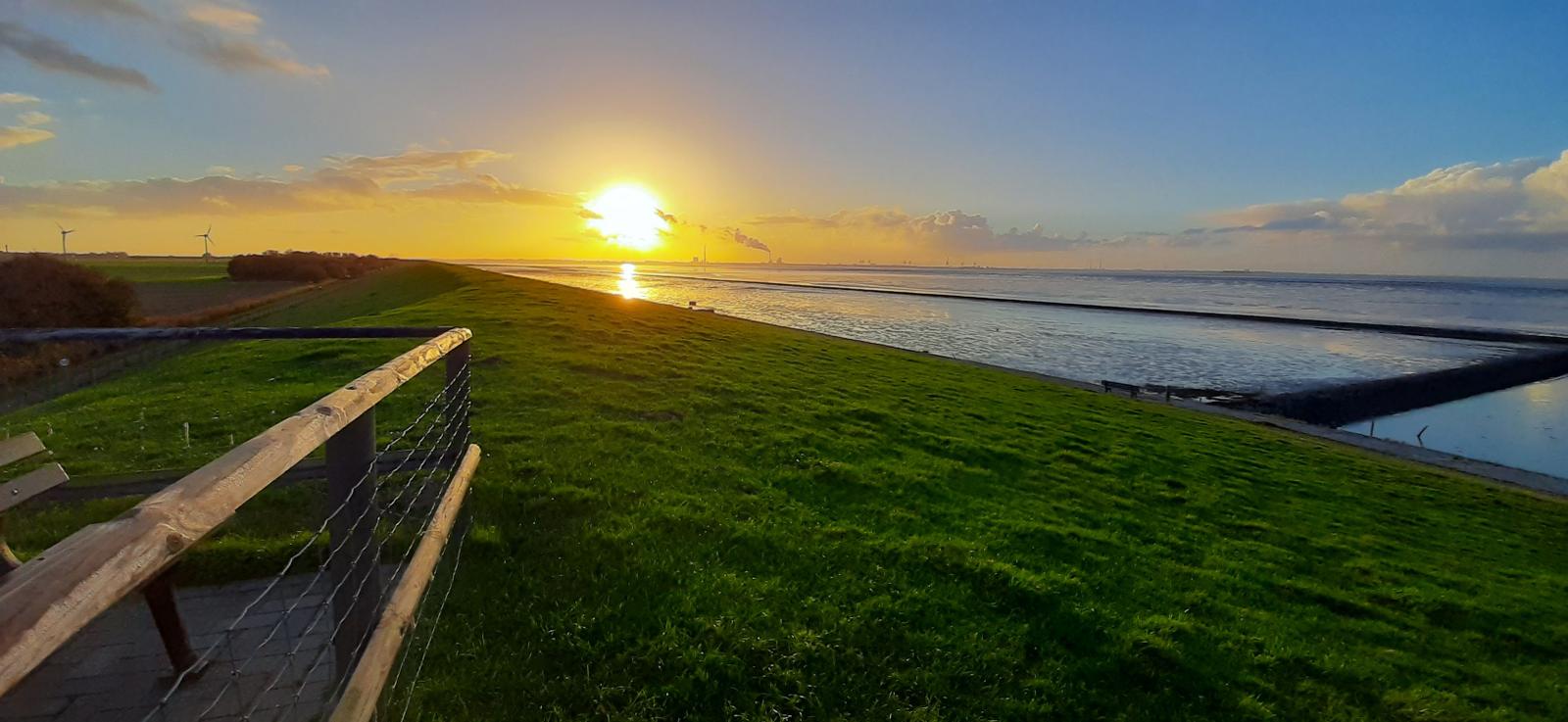 Sonnenuntergang über dem Wasser mit grünem Hang und Holzgeländer
