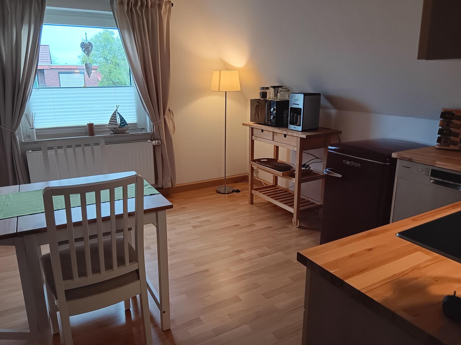 Essbereich mit Tisch, Stühlen und Küchenzeile. Fenster mit Vorhängen und Lampe.