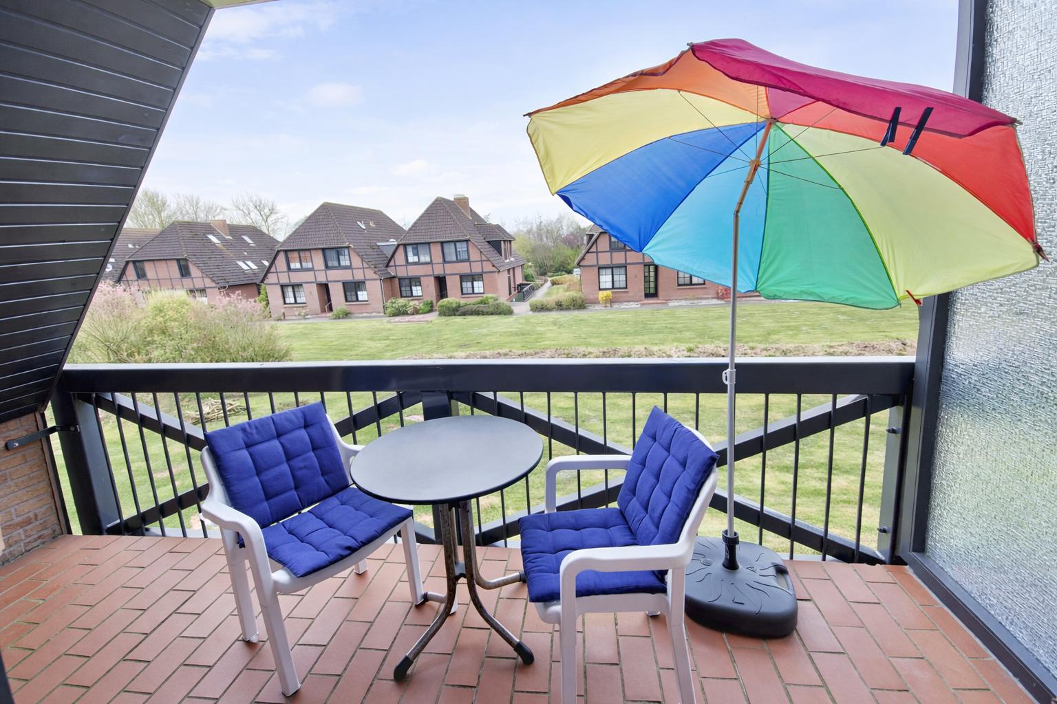 Terrasse mit Tisch, zwei Stühlen und buntem Sonnenschirm. Blick auf Grünfläche und Häuser.