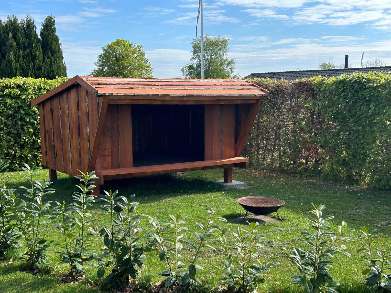 Holzschuppen mit Sitzbank im Garten, daneben Grill, umgeben von Hecken und Gras.