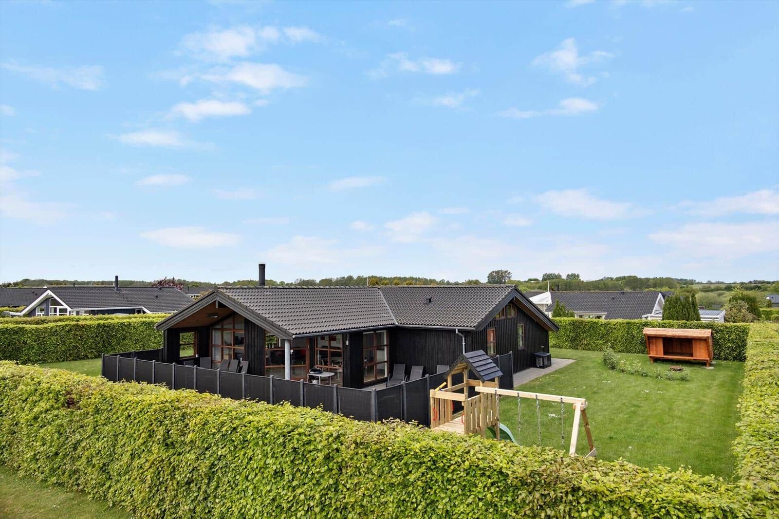 Holzhaus mit Garten, Spielplatz und Grillbereich. Umzäunt und von Hecken umgeben.