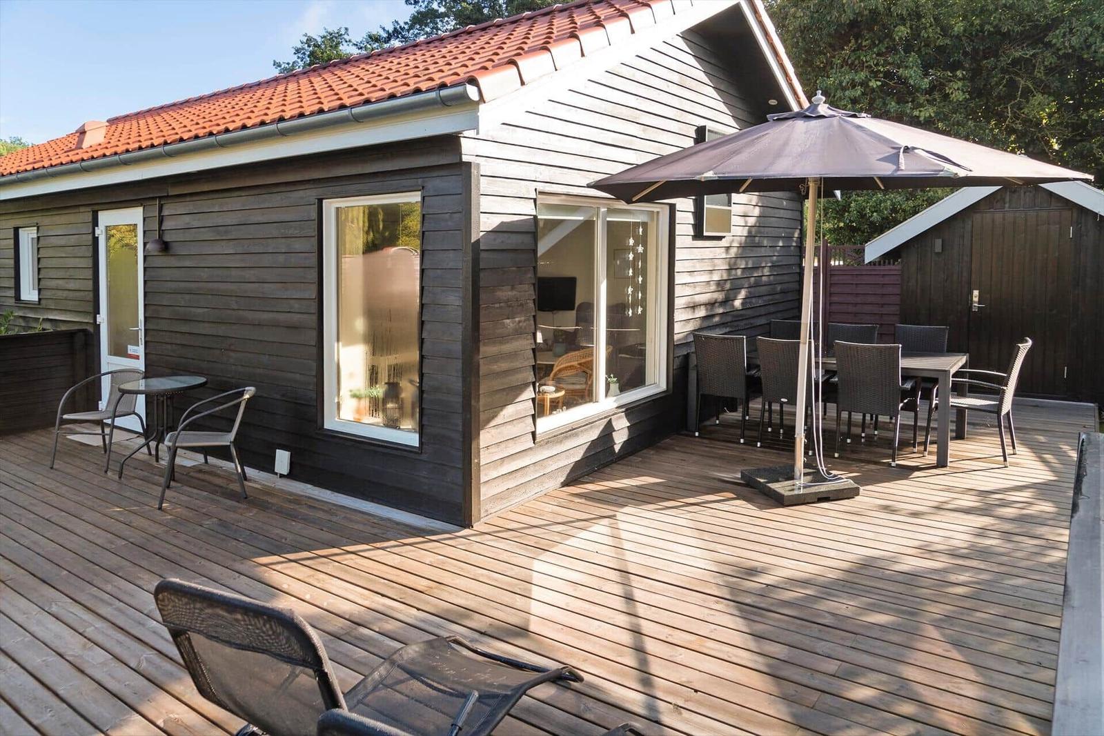 House with wooden terrace, seating, and umbrella. Large windows show interior.