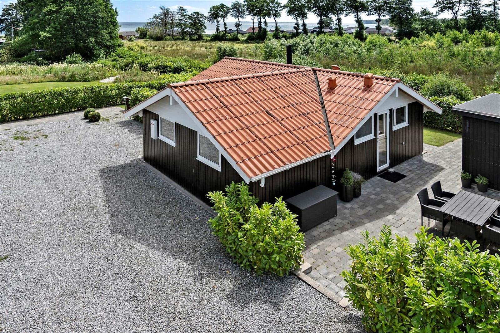 A dark brown holiday home with a red tiled roof, terrace, and garden. View of the water in the background.