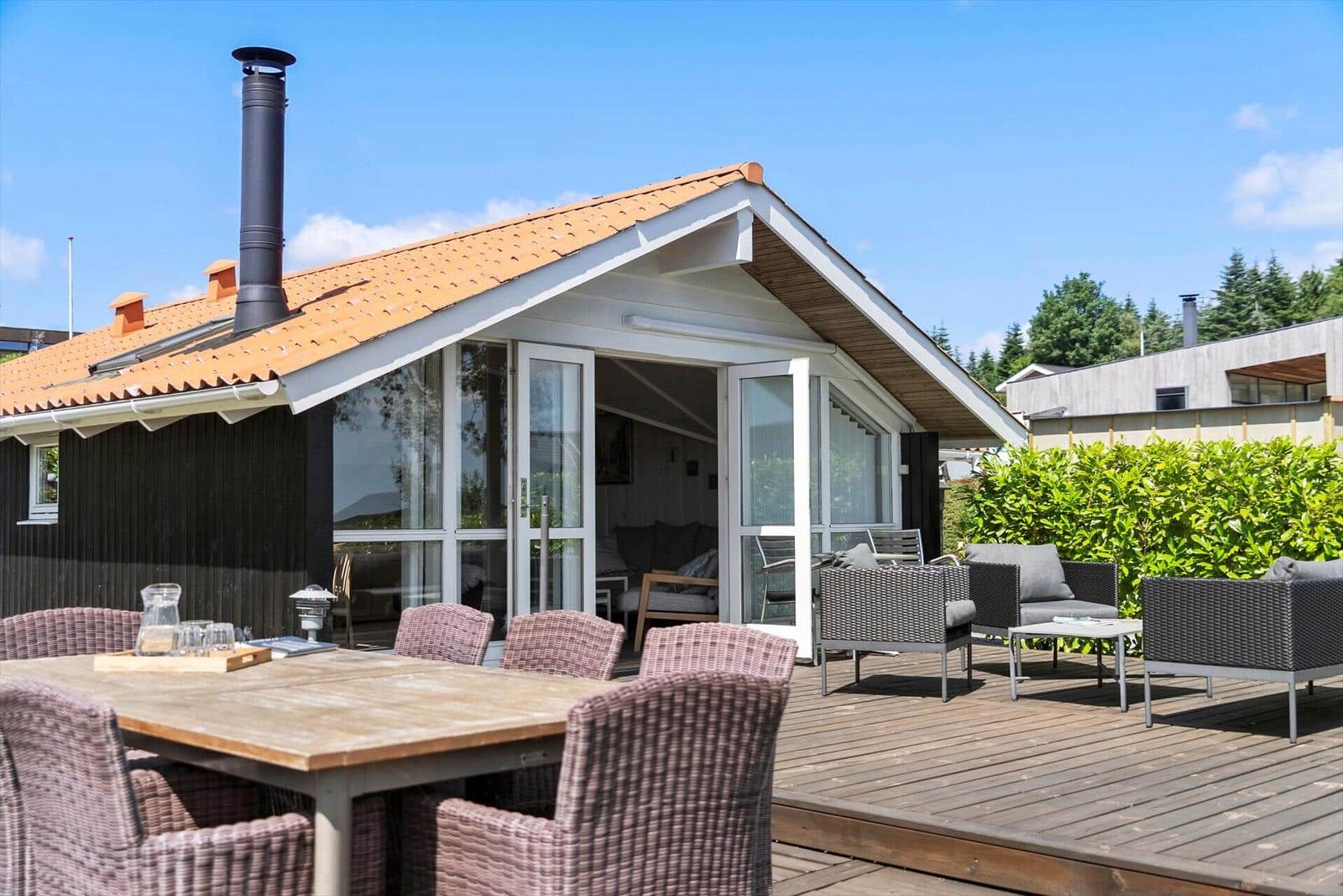 House with terrace, garden furniture, and chimney. Large window front. Wooden terrace with table and chairs.