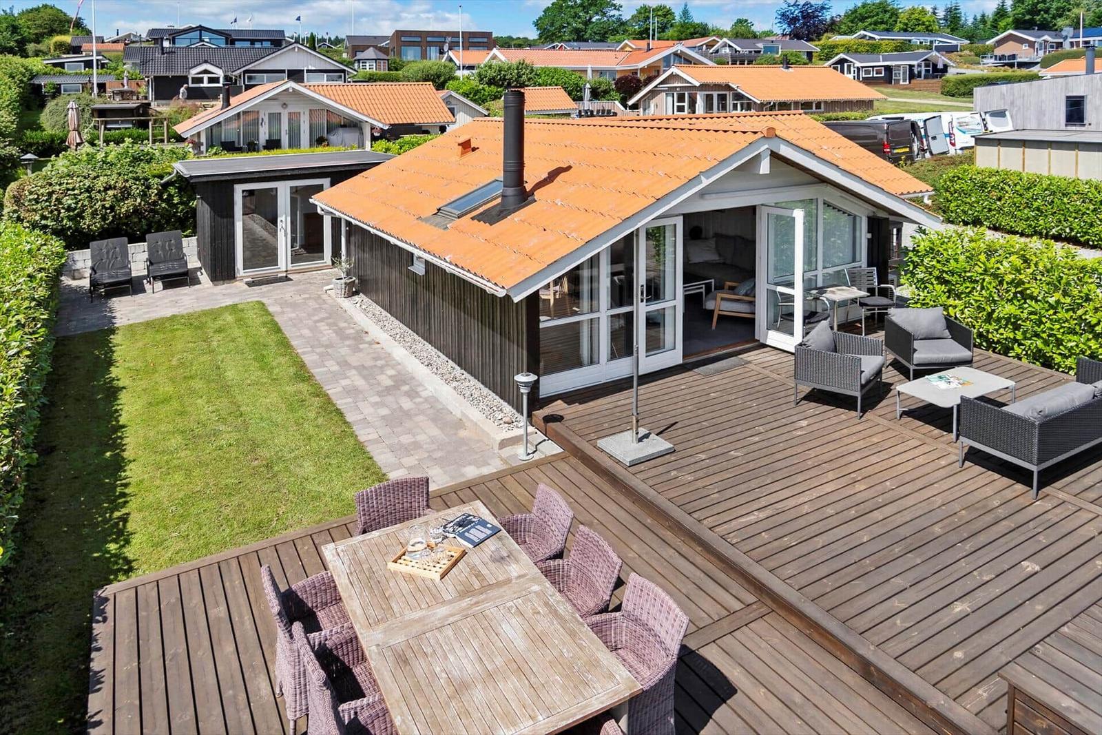 House with wooden terrace, garden, and seating areas. Large glass front opens to the outside.