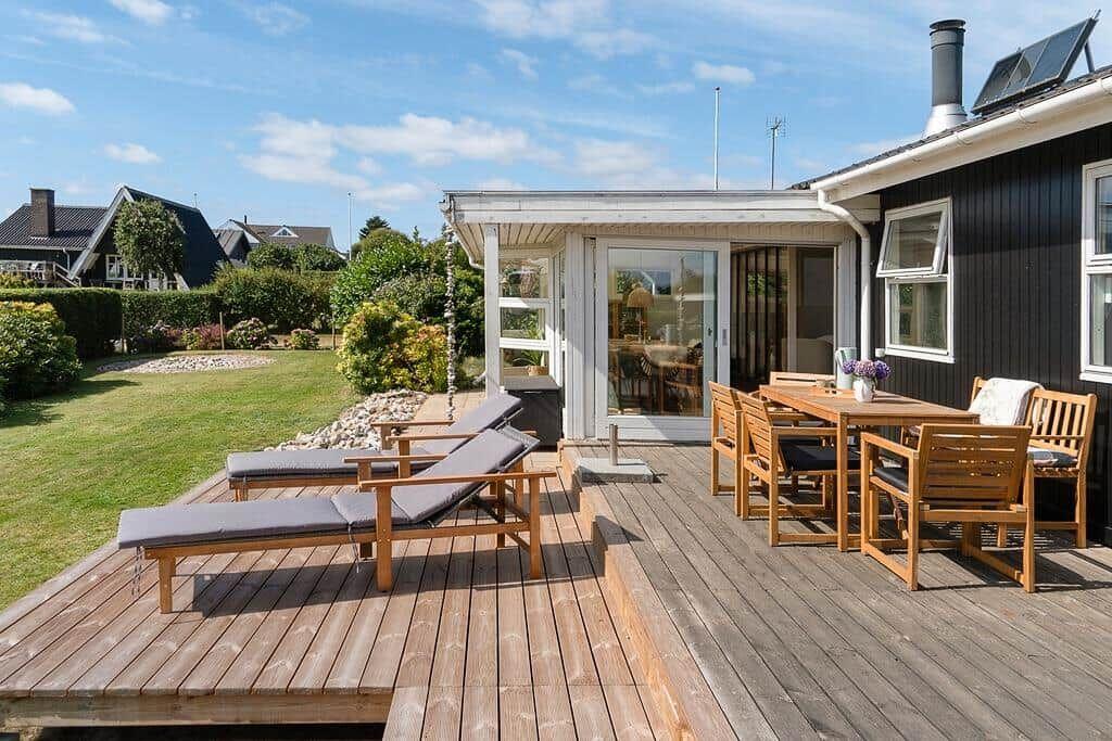 Terrasse mit Liegestühlen und Tisch. Hinterhof mit Rasen und Hecke. Haus mit Fenstern und Dach.
