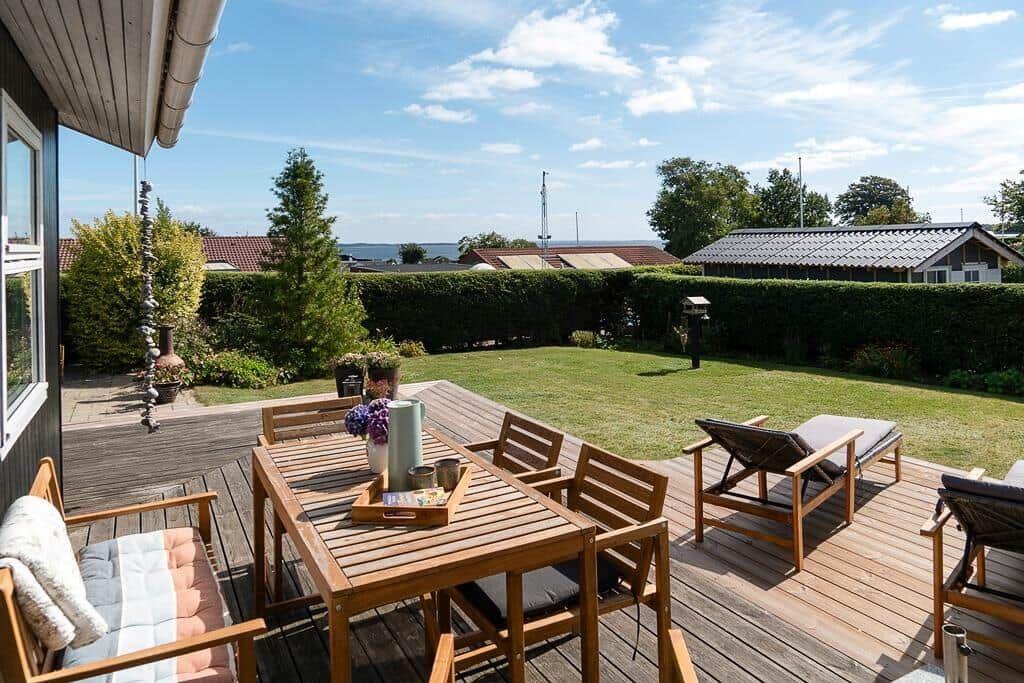 Terrasse mit Holztisch, Stühlen und Liegen. Grünfläche und Hecke im Hintergrund.