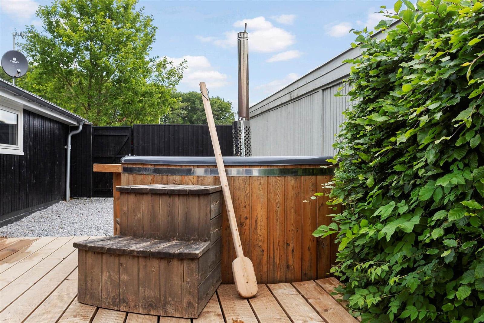 Holzwhirlpool mit Stufen und Schaufel auf Terrasse. Umgeben von Grün und Hütten.