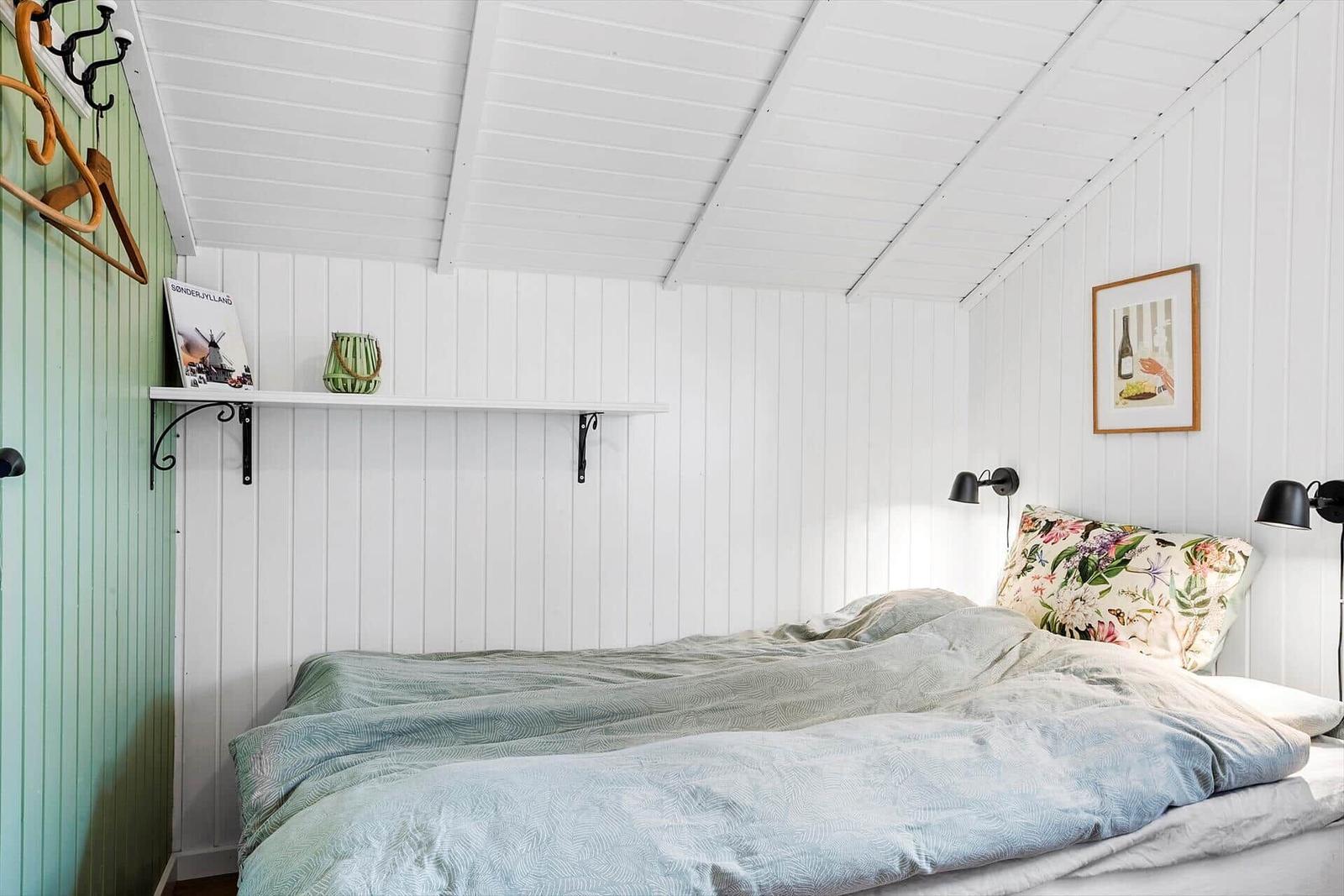 Schlafzimmer mit Bett, weißen Wänden und Decke, Wandregal und Bild an der Wand.