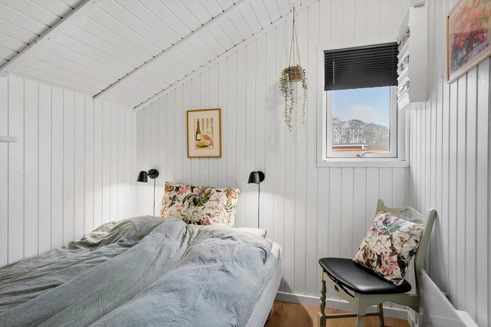 Schlafzimmer mit Bett, Stuhl und Fenster. Wände und Decke weiß verkleidet.
