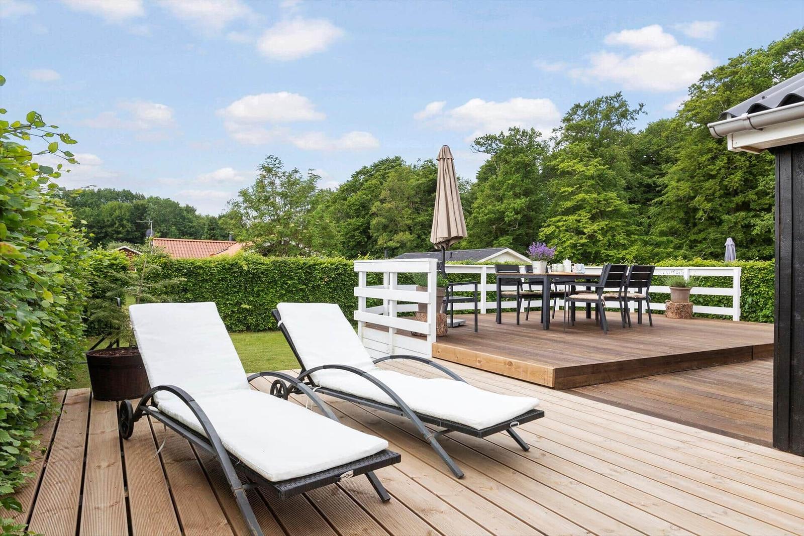 Terrasse mit zwei Liegestühlen, Esstisch und Grillplatz. Blick auf grüne Bäume und Himmel.
