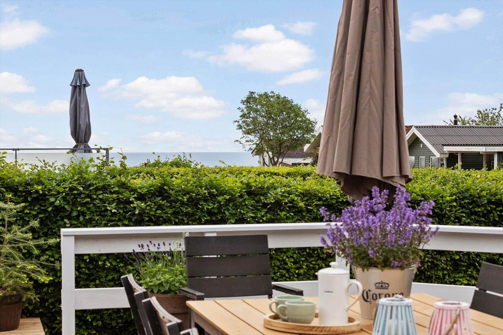 Terrasse mit Tisch, Stühlen und Sonnenschirmen. Blick auf Meer und grüne Hecke.