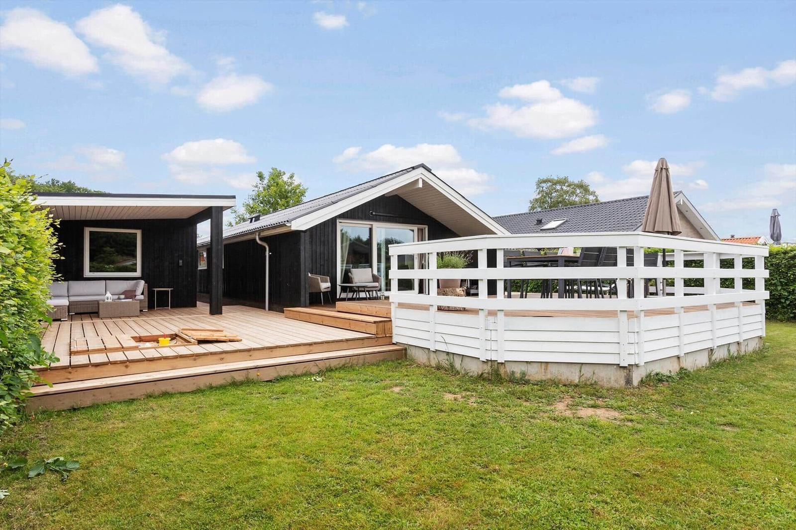 Holzterrasse mit Sitzgelegenheiten und weißem Geländer. Modernes Haus mit großem Fenster.