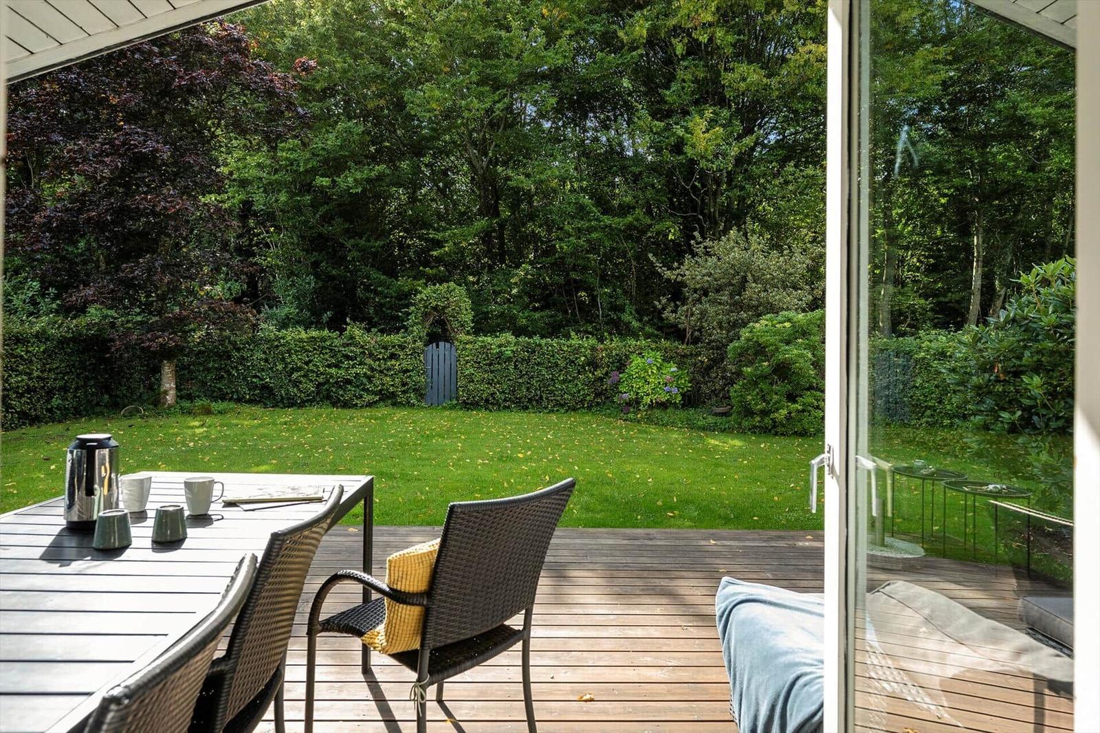 Terrasse mit Tisch, Stühlen und Blick auf grünen Garten mit Hecke und Bäumen.