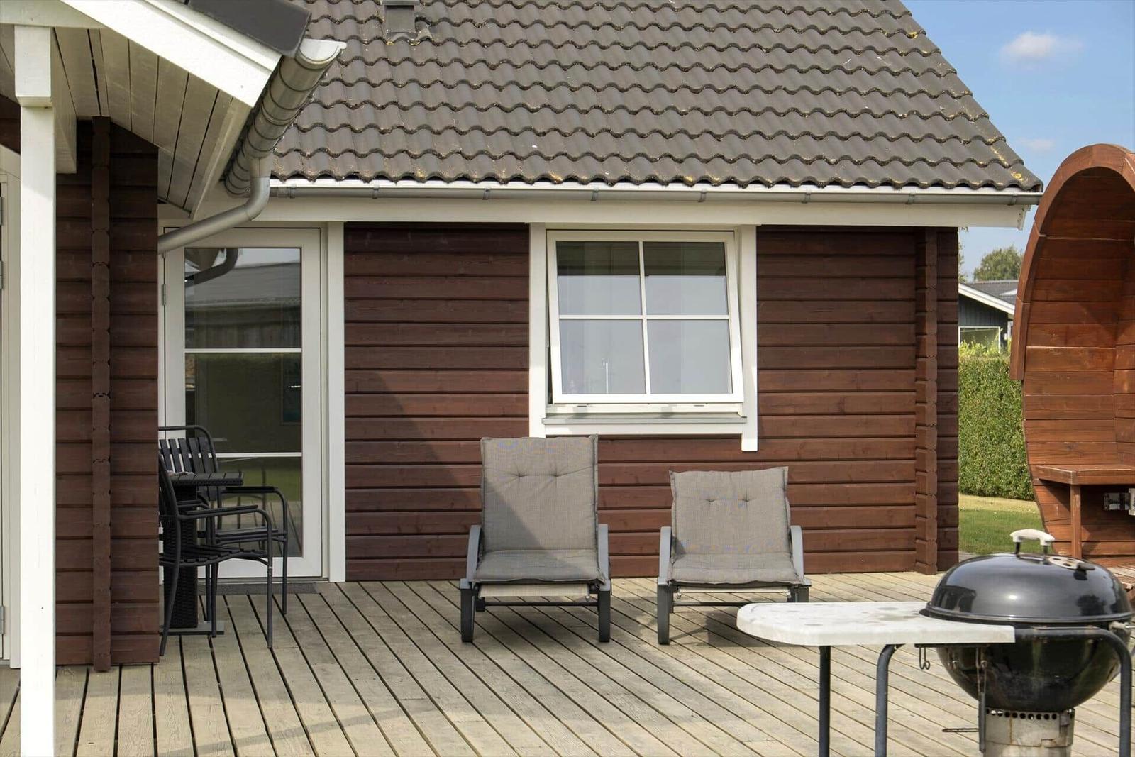 Terrasse mit zwei Liegestühlen, Grill und Tisch vor einem Holzhaus mit Dach und Fenster.