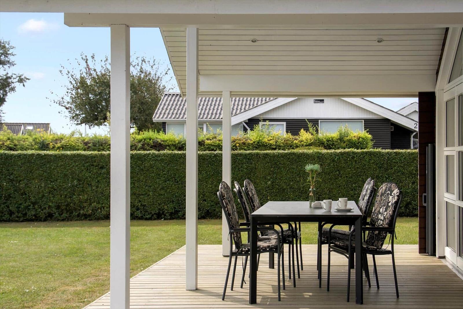 Terrasse mit Tisch und Stühlen unter weißem Dach. Grünfläche und Hecke im Hintergrund.