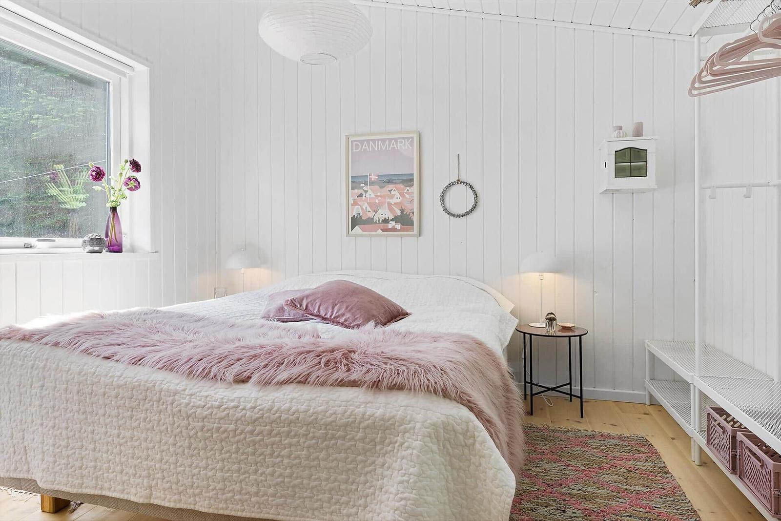 Schlafzimmer mit weißer Holzwand, Bett mit rosa Decke und Kissen, Fenster mit Blumen und Wandbild.