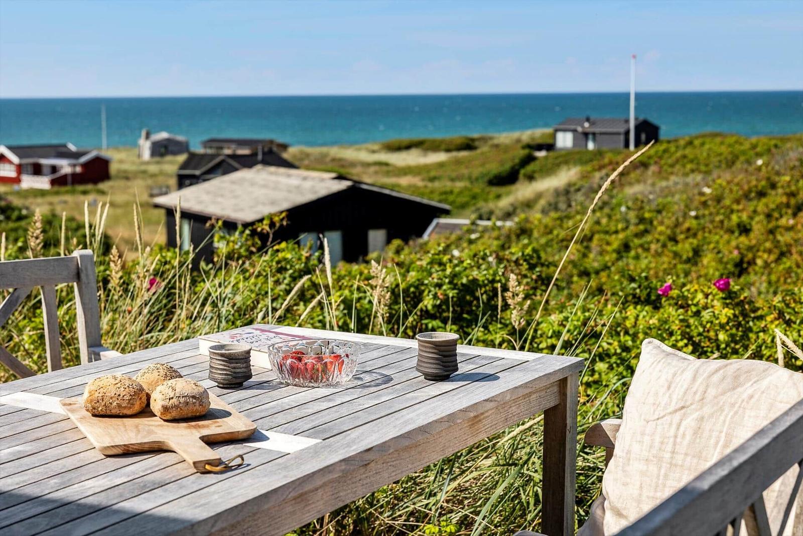Holztisch mit Brot, Beeren und Tassen auf Terrasse mit Blick auf das Meer und Gras.