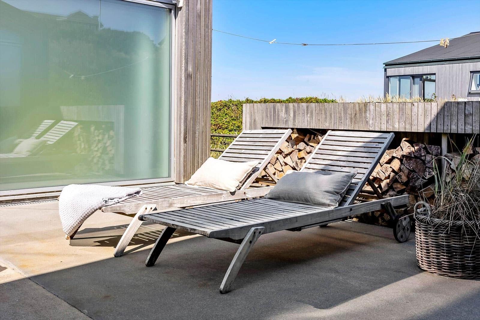 Zwei Holzliegestühle mit Kissen stehen auf einer Terrasse neben einem Holzstapel.