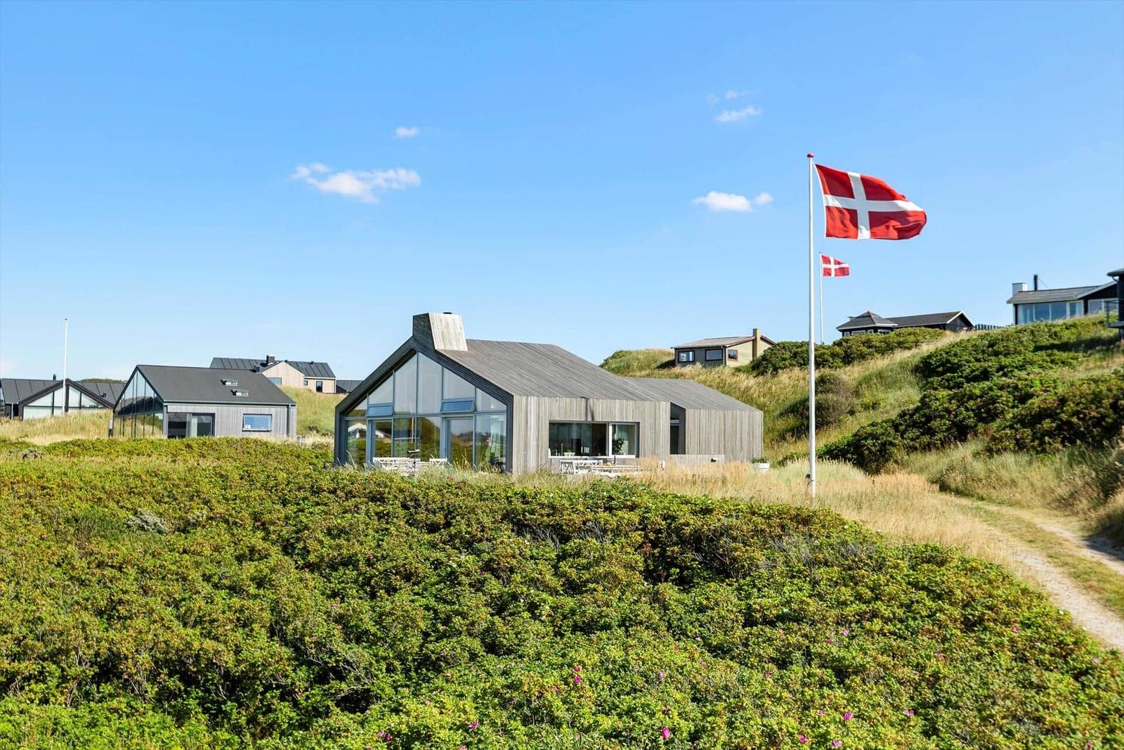 Moderne Villa mit Glasfassade und dänischer Flagge auf grüner Dünenlandschaft.