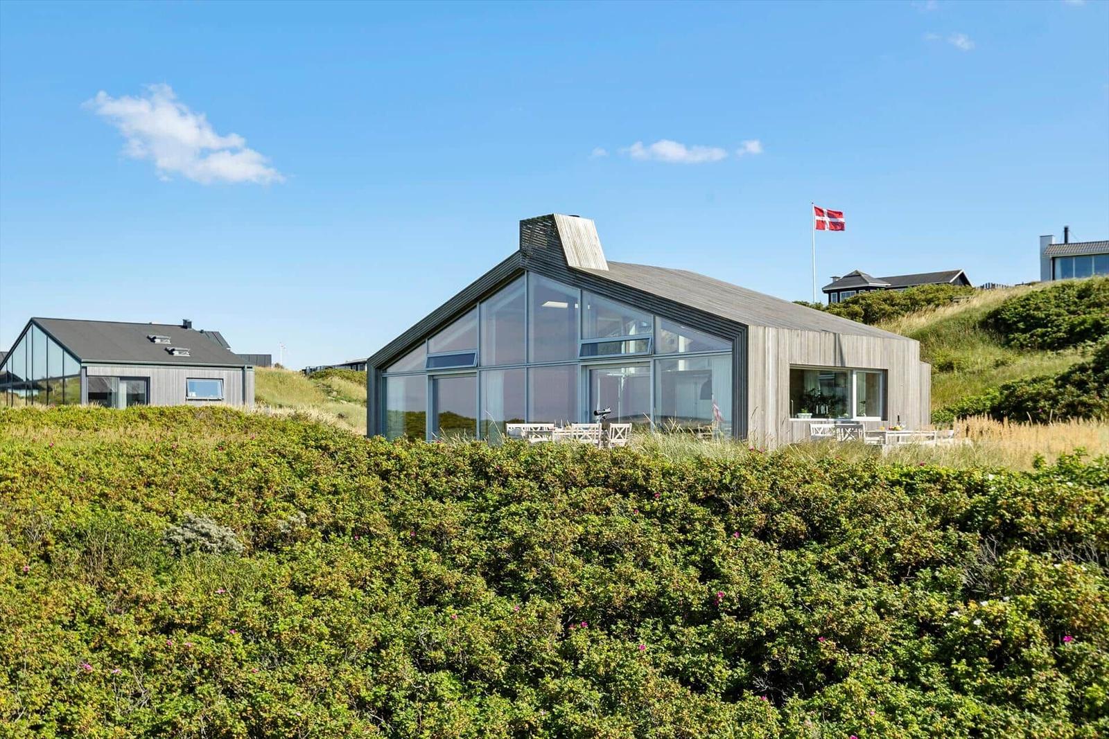 Moderne Villa mit Glasfassade und Terrasse auf grüner Dünenlage. Dänische Flagge weht oben.