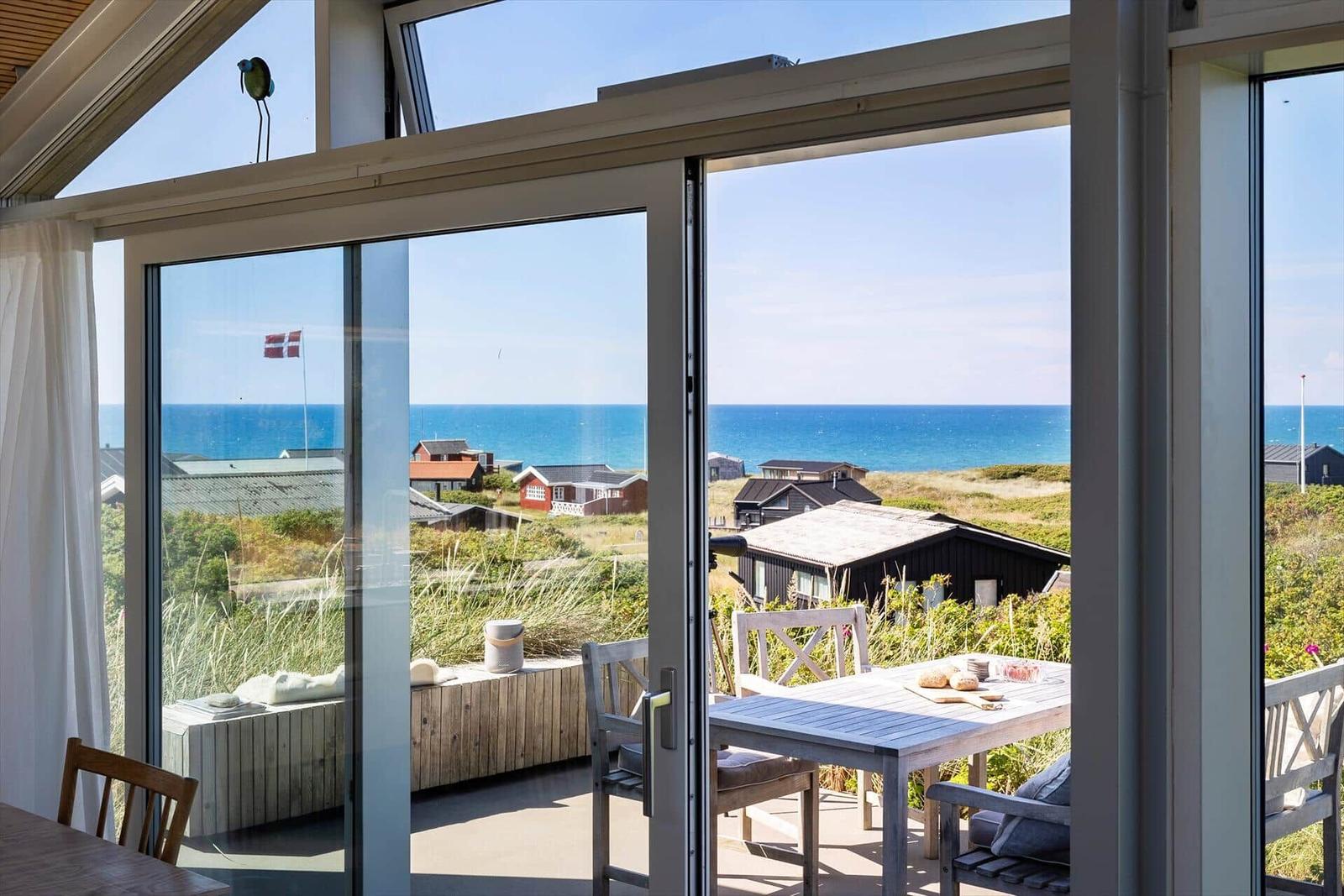 Grosses Fenster mit Blick aufs Meer und Strandhaus. Terrasse mit Tisch und Stühlen.