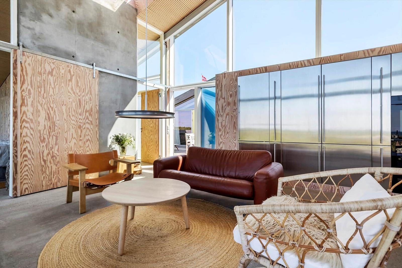 Wohnzimmer mit Ledersofa, Rattan-Sessel und Holztisch. Große Fenster und Holzwände.